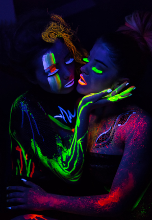 Sexy lesbian fashion models in uv neon light with fluorescent glowing Body Art make-up hugging kissing. Low key dark image. Soft focus image.の写真素材