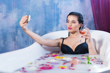 Sexy brunette woman taking selfie with glass of champagne having a hot milk bath with flower Petals. she is wearing black sexual lingerieの写真素材