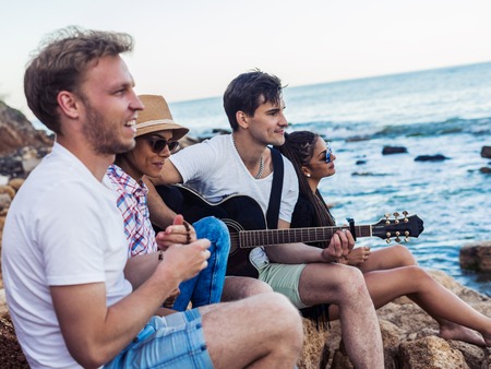 friends sitting on stones on beach. man is playing guitar.の写真素材