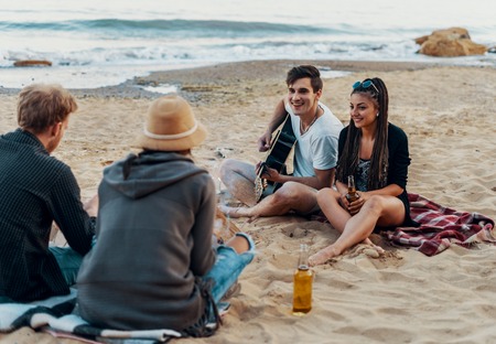 friends sitting on stones on beach. man is playing guitar.の写真素材