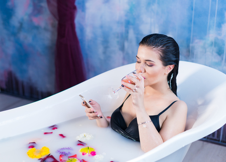 Sexy brunette woman is sitting in bath and chatting on smartphone
