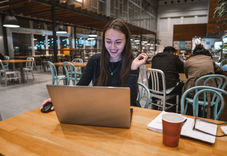 Young, beautiful girl working with laptop and drinking coffee at a wooden tableの写真素材
