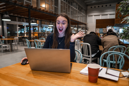 Young, beautiful girl working with laptop and drinking coffee at a wooden tableの写真素材