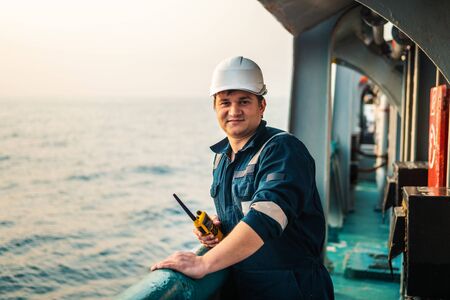 Marine Deck Officer or Chief mate on deck of offshore vessel or ship , wearing PPE personal protective equipment - helmet, coverall. He holds VHF walkie-talkie radio in hands.の写真素材