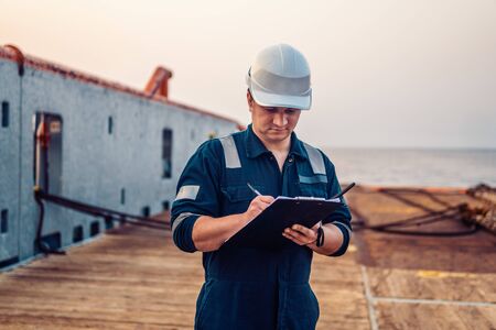 Deck Officer on deck of offshore vessel or ship , wearing PPE personal protective equipment. He fills checklist. Paperwork at seaの写真素材