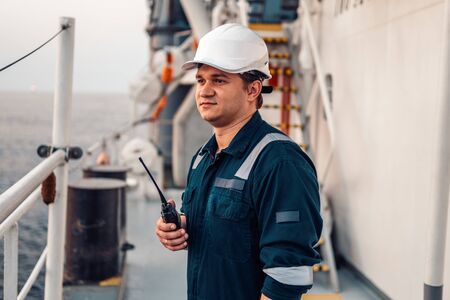 Marine Deck Officer or Chief mate on deck of offshore vessel or ship , wearing PPE personal protective equipment - helmet, coverall. He holds VHF walkie-talkie radio in hands.の写真素材