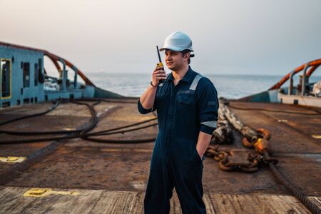 Marine Deck Officer or Chief mate on deck of offshore vessel or ship , wearing PPE personal protective equipment - helmet, coverall. He holds VHF walkie-talkie radio in hands.の写真素材