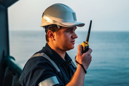 Marine Deck Officer or Chief mate on deck of offshore vessel or ship , wearing PPE personal protective equipment - helmet, coverall. He holds VHF walkie-talkie radio in hands.の写真素材
