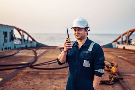 Marine Deck Officer or Chief mate on deck of offshore vessel or ship , wearing PPE personal protective equipment - helmet, coverall. He holds VHF walkie-talkie radio in hands.の写真素材