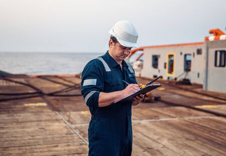 Deck Officer on deck of offshore vessel or ship , wearing PPE personal protective equipment. He fills checklist. Paperwork at seaの写真素材