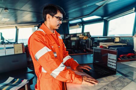 Filipino deck Officer on bridge of vessel or ship. He is using laptop, electronic paperwork at seaの写真素材