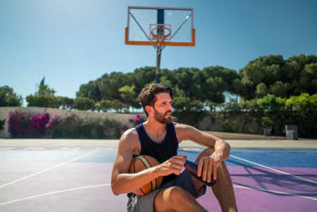 tired basketball player after the match, sits on the basketball field and drinks waterの写真素材