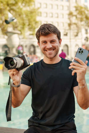 Cheerful young guy sitting near fountain with modern photo camera and smartphoneの写真素材