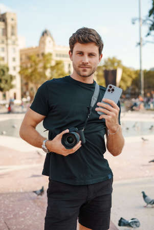 Self assured guy with camera and smartphone standing on city squareの写真素材