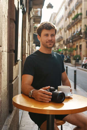 Smiling guy resting at table in outdoor cafe with coffee cup and camera