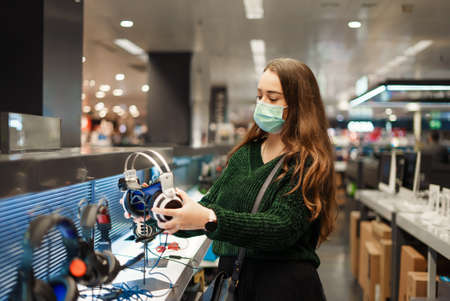 Young woman in medical face mask choosing modern headphones, shopping during Covid19 outbreakの写真素材