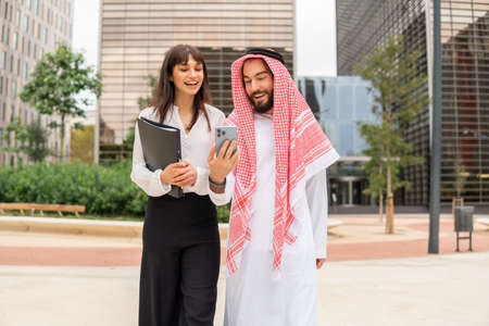 Smiling Arabian businessman and his female assistant watching funny videos on smartphone outdoorsの写真素材