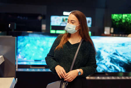 Young woman in medical face mask pensively examines variety of tv sets on display in tech storeの写真素材