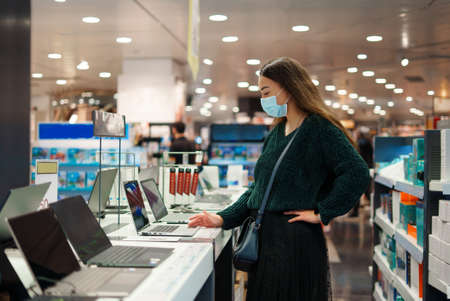 Focused young woman in face mask examines laptop on display in tech store department