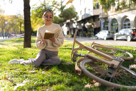 Smiling woman reading book on grass near eco friendly bicycleの写真素材