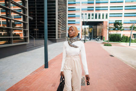 Stylish black Muslim woman with bag observing high buildingsの写真素材