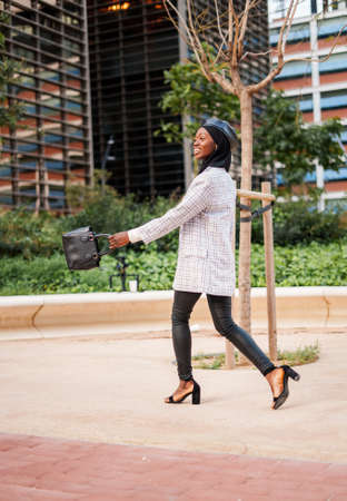 Cheerful black lady in stylish wear walking in city parkの写真素材
