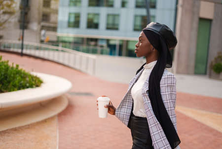 Side view of African American female in stylish outfit and Islamic headscarf drinking takeaway coffee in urban parkの写真素材