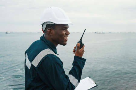 Happy African American man in blue uniform and hardhat with clipboard smiling and talking to colleagues on VHF walkie talkie against rippling sea and cloudy sky during work on shipの写真素材