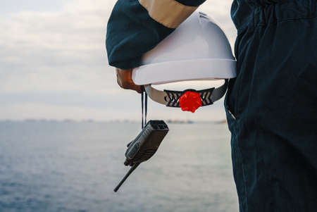 Unrecognizable seaman in uniform with white hardhat and VHF walkie talking admiring sea and overcast sky while working on ship in daytimeの写真素材