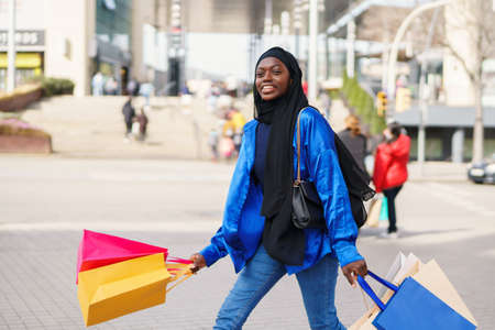Positive black female in casual clothes and hijab looking away with smile and swinging colorful paper bags while walking on paved sidewalk after shopping in weekend in cityの写真素材