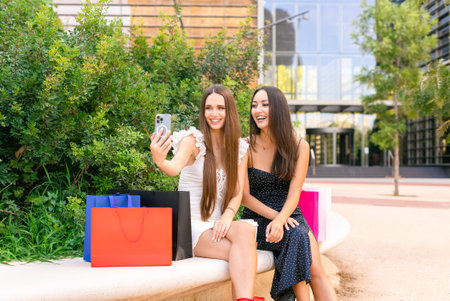 Trendy girlfriends in dresses shooting selfie on city benchの写真素材