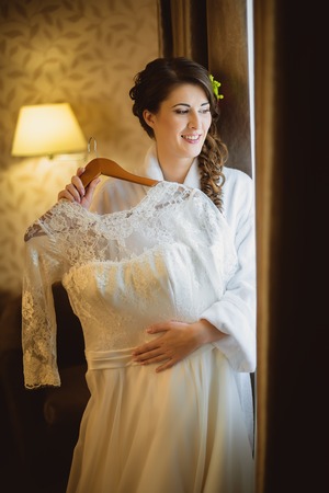 Bride holds a wedding dress standing near a windowの写真素材