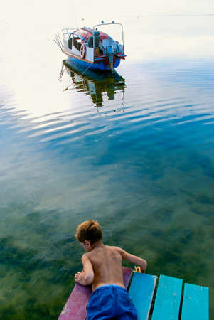 Boy lying on a pier looking in the water standing in the background boats in the bay.の写真素材
