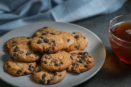 A cup of hot black tea in a glass cup and a gray ceramic plate with cookies with chocolate chips on a gray ceramic counter top with a kitchen towel.の写真素材