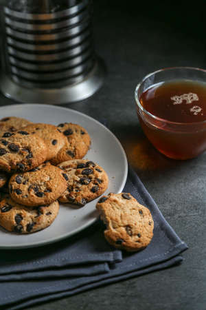A cup of hot black tea in a glass cup and a teapot press and a gray ceramic plate with cookies with chocolate chips on a gray ceramic counter top with a gray linen napkin.の写真素材