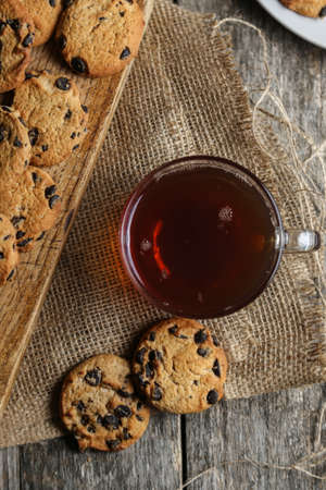 A cup of hot black tea in a glass cup and a gray ceramic plate with cookies with chocolate chips on a wooden tabletop with a board and burlap.の写真素材