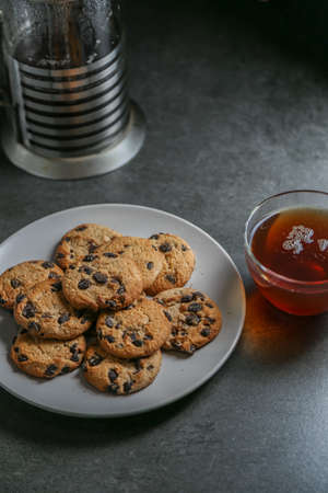 A cup of hot black tea in a glass cup and a teapot press and a gray ceramic plate with cookies with chocolate chips on a gray ceramic tabletop.の写真素材