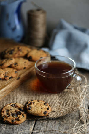A cup of hot black tea in a glass cup and a teapot press and a gray ceramic plate with cookies with chocolate chips on a wooden tabletop with a board and sackcloth.の写真素材