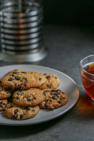 A cup of hot black tea in a glass cup and a teapot press and a gray ceramic plate with cookies with chocolate chips on a gray ceramic tabletop.の写真素材