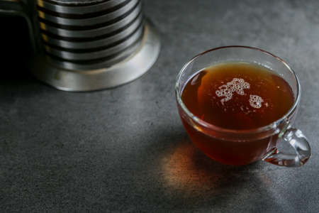 A cup of hot black tea in a glass cup and a kettle press on a gray ceramic countertop.の写真素材