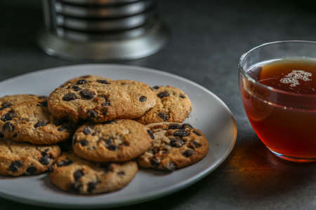 A cup of hot black tea in a glass cup and a teapot press and a gray ceramic plate with cookies with chocolate chips on a gray ceramic tabletop.の写真素材