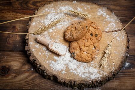 Oatmeal cakes with raisins, flour and spikelets of wheat on a wooden brown surfaceの写真素材