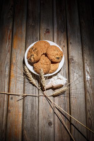 Oatmeal cakes with raisins, flour and spikelets of wheat on a wooden brown surfaceの写真素材