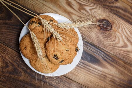 Oatmeal cakes with raisins, flour and spikelets of wheat on a wooden brown surfaceの写真素材