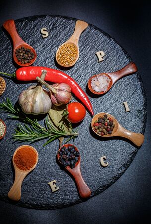 Spices in wooden spoons and cherry tomatoes, chili peppers, garlic and herbs for cooking on a black surface and black backgroundの写真素材