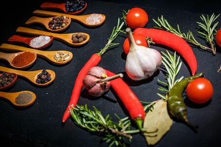 Spices in wooden spoons and cherry tomatoes, chili peppers, garlic and herbs for cooking on a black surface and black backgroundの写真素材