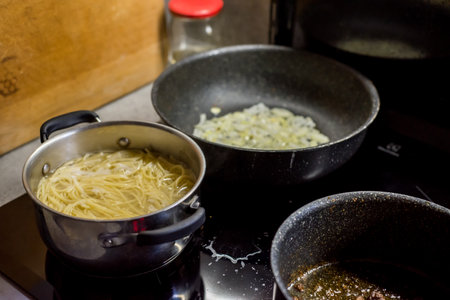 Pasta boils in metal pot, nearby pan holds sliced onion frying, second pan contains meat searing in sauce. All three vessels rest on black stovetop, showing busy cooking in home kitchen setup.の写真素材