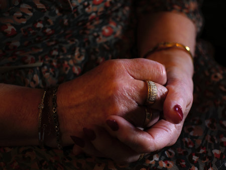 Close up of 70s woman clasped hands, resting on her knees. She has bracelets, rings and red nailsの写真素材