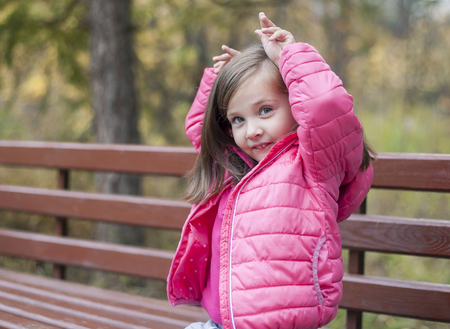 Little pretty girl in a pink coat sitting on a wood bench at the park in autumn. Emotional portrait. Childhood concept. Caucasian. Child with brunette long hair. Leisure, relaxation, lifestyleの写真素材