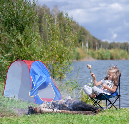 Little girl sitting near campfire. Girl roasting bread over campfire. Tourist relaxing near campfire. Rest at the lakeの写真素材